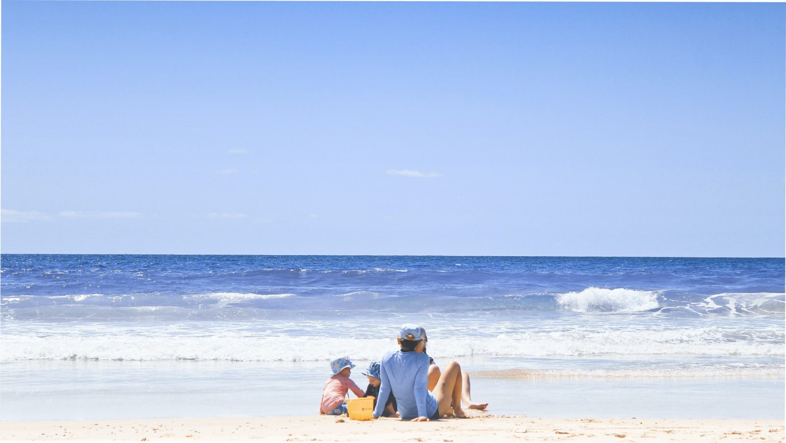 Two white children, wearing blue hats, and a white man, wearing a shirt and blue hat, both sitting on the sand in front of the sea