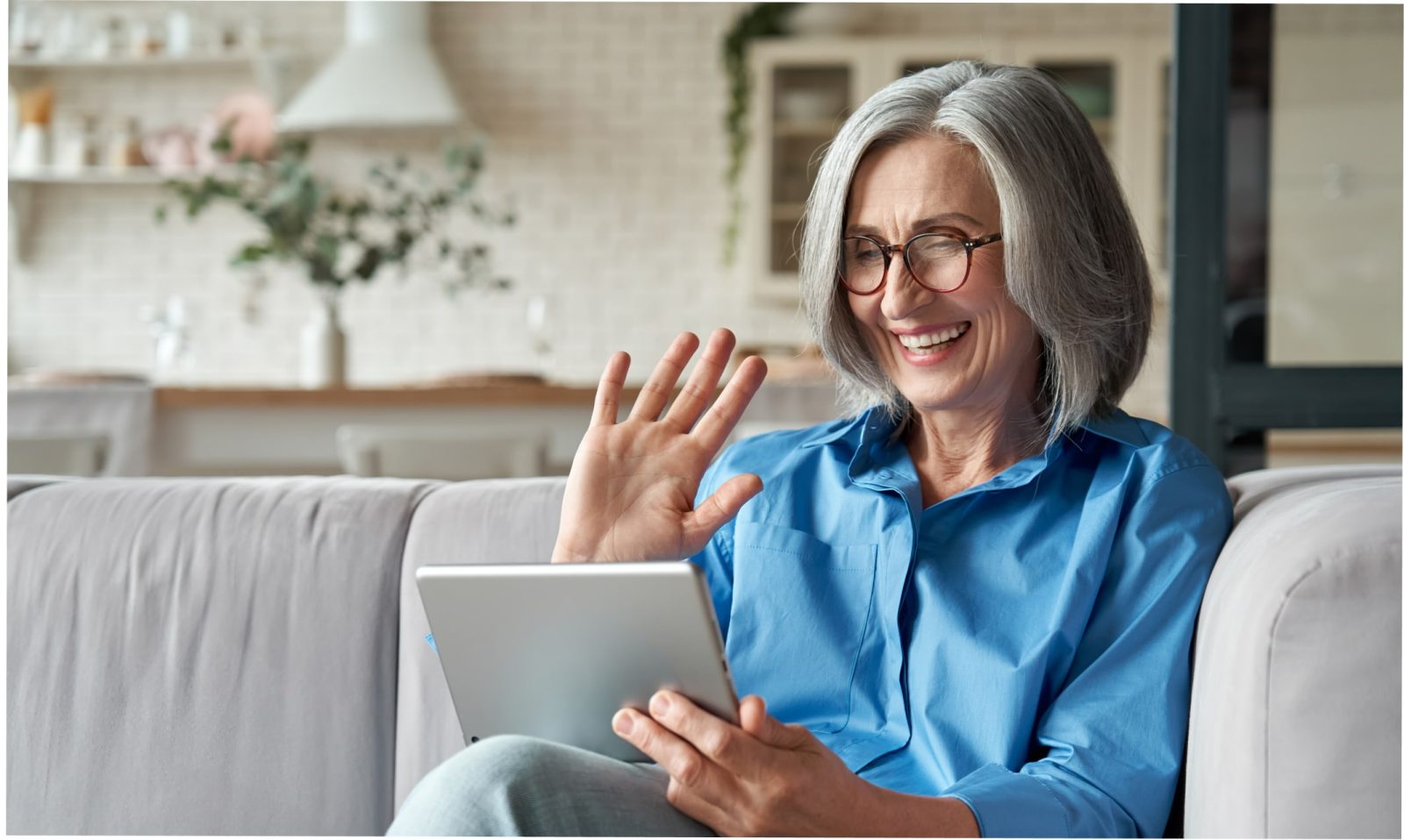 White woman with white hair, wearing brown glasses, a blue shirt, sitting on a gray sofa, while holding a gray tablet with her left hand and waving with her right hand, smiling at the screen