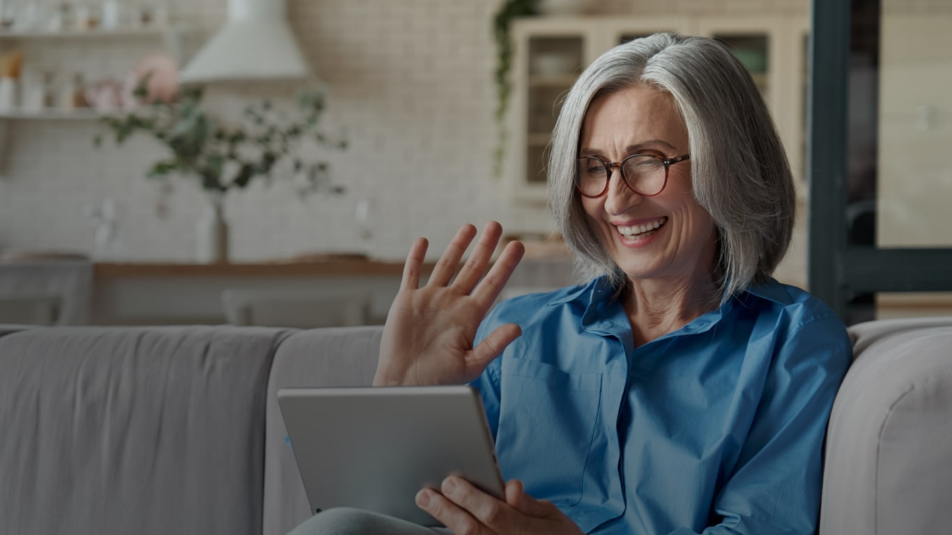 White woman with white hair, wearing brown glasses, a blue shirt, sitting on a gray sofa, while holding a gray tablet with her left hand and waving with her right hand, smiling at the screen