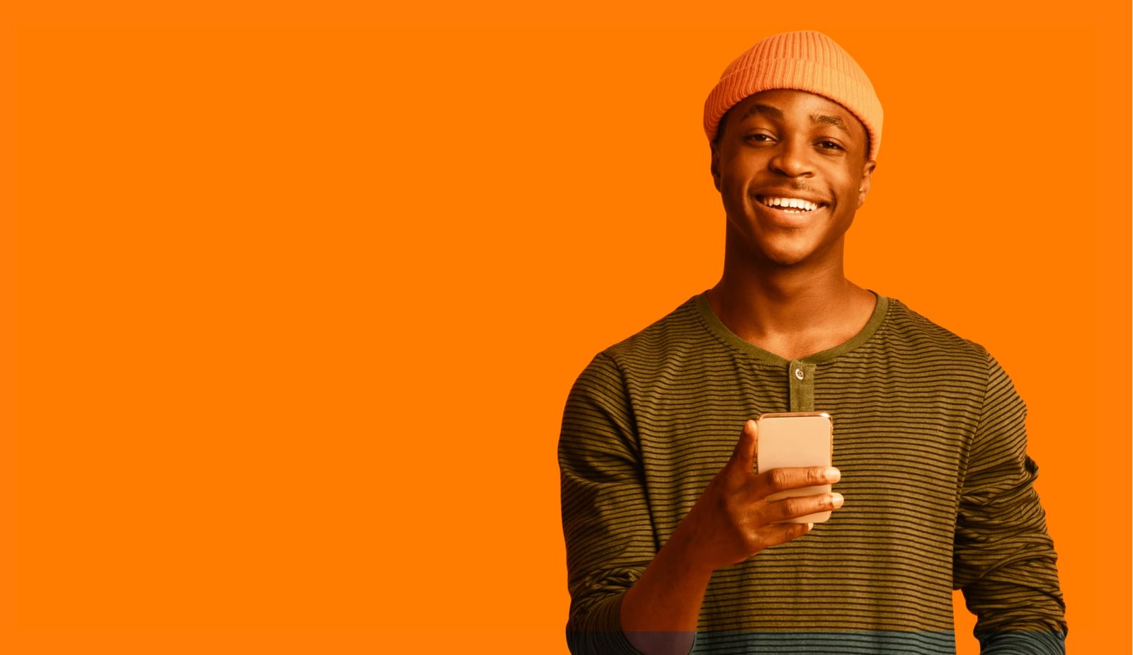 Black man standing, wearing an orange cap and green striped t-shirt, while holding a white cell phone with his right hand and posing smiling