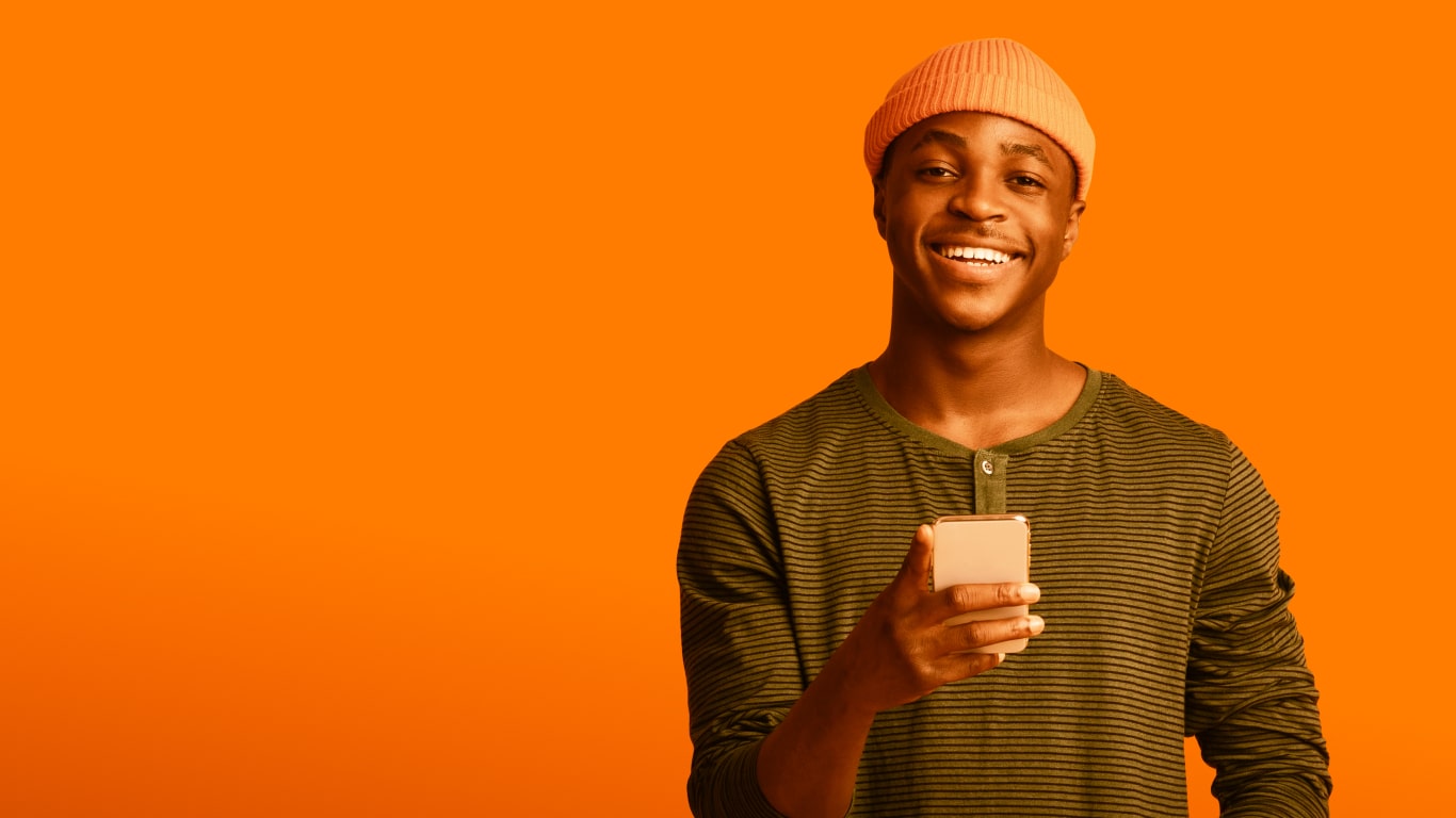 Black man standing, wearing an orange cap and green striped t-shirt, while holding a white cell phone with his right hand and posing smiling