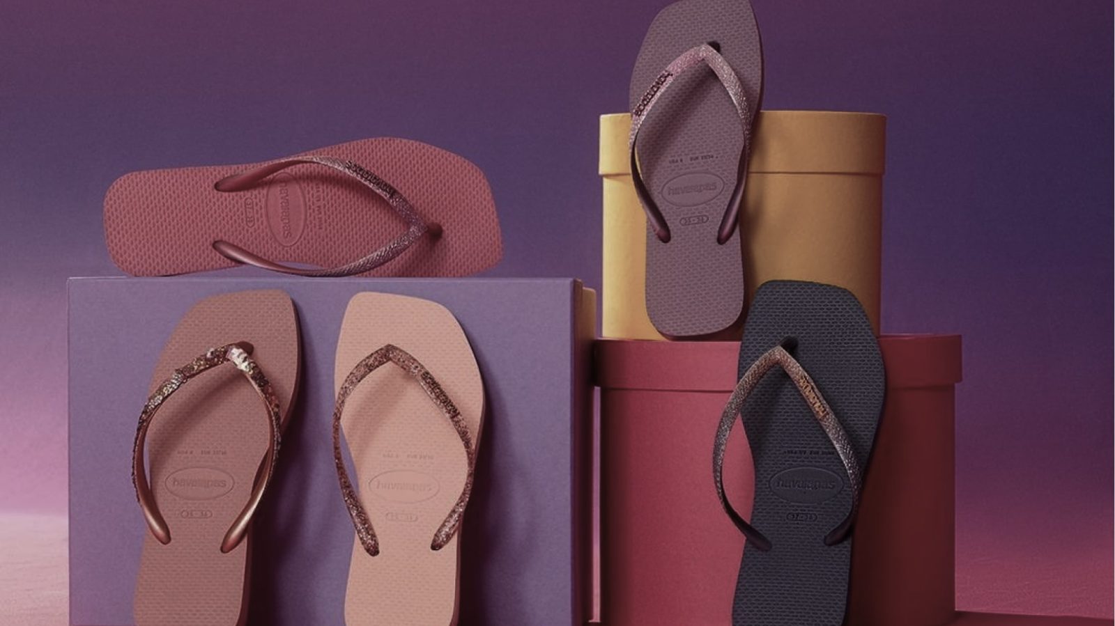 Five Havaianas flip-flops in different colors, stacked on boxes, a purple box, a pink box and a yellow box, in front of a plain purple background