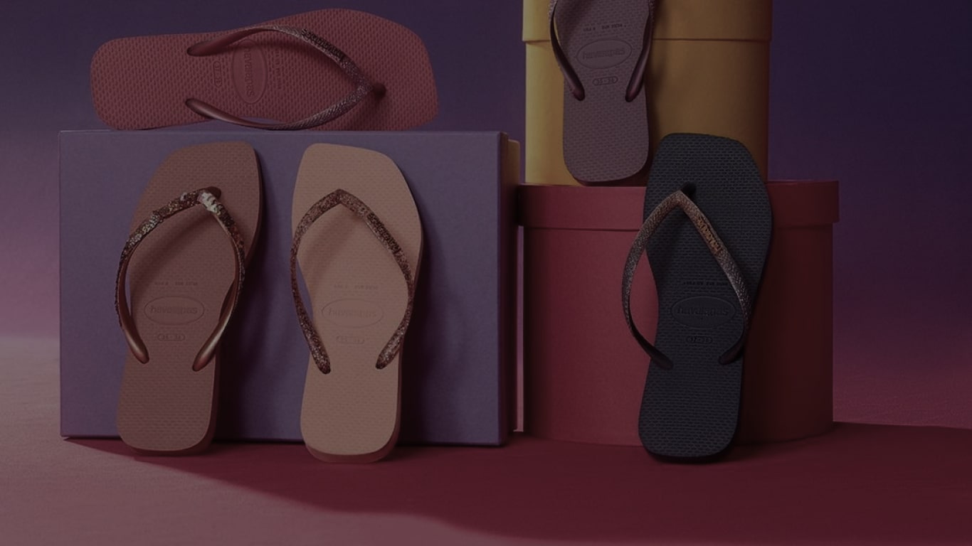 Five Havaianas flip-flops in different colors, stacked on boxes, a purple box, a pink box and a yellow box, in front of a plain purple background