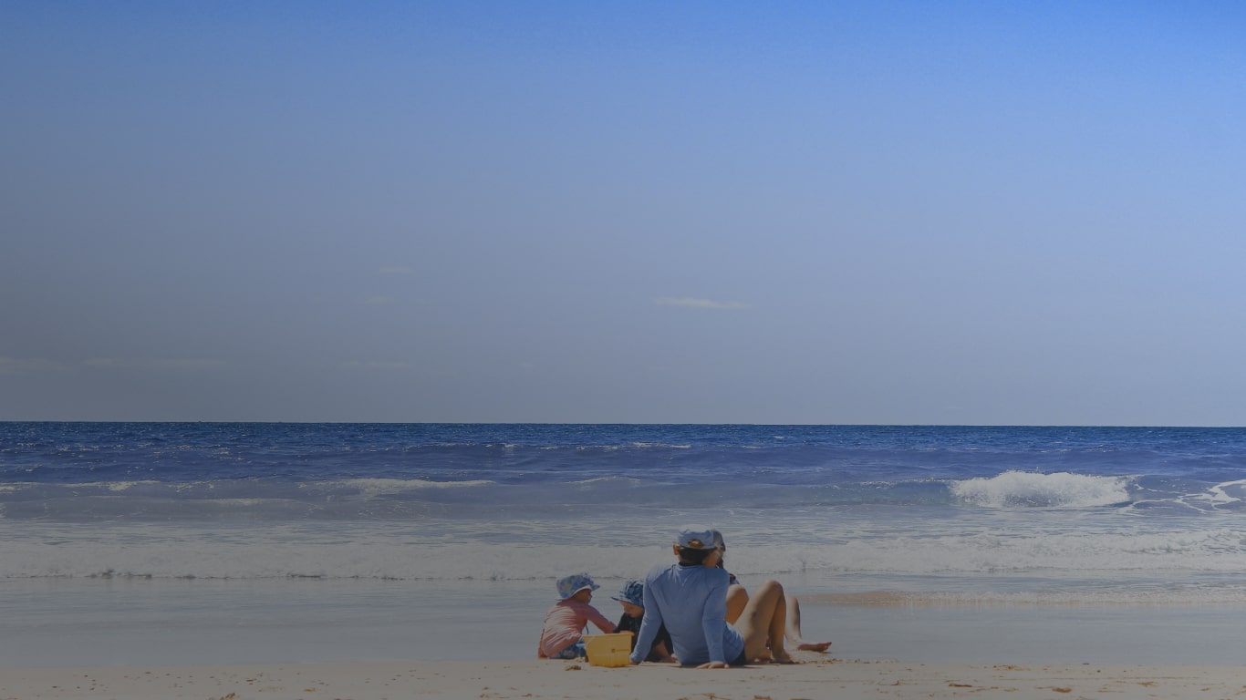 Duas crianças brancas, vestindo chapéus azuis, e um homem branco, vestindo camiseta de manga longa e chapéu azul, ambos sentados na areia de frente para o mar